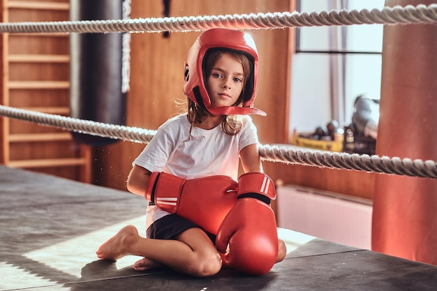 Una joven deportiva está sentada en un anillo soleado usando su equipo: casco y guantes de boxeo.