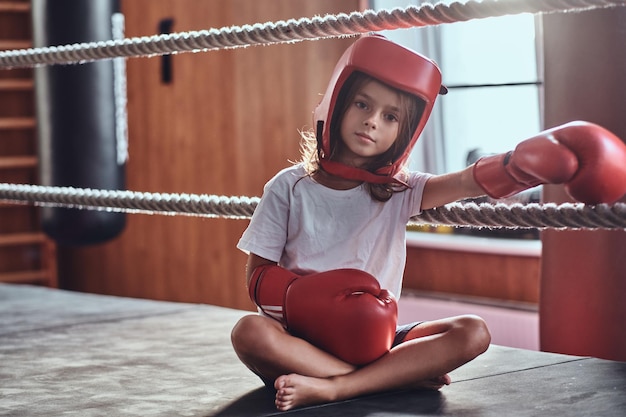 Una joven deportiva está sentada en un anillo soleado usando su equipo: casco y guantes de boxeo.