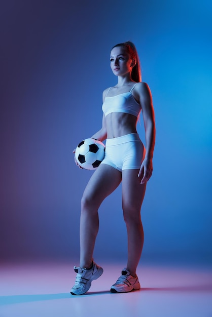 Joven deportiva en ropa deportiva blanca entrenando posando con pelota de fútbol aislada sobre gradiente