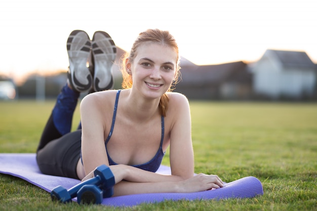 Joven deportiva en ropa deportiva tendido sobre la colchoneta de entrenamiento antes de hacer ejercicios en el campo al amanecer.