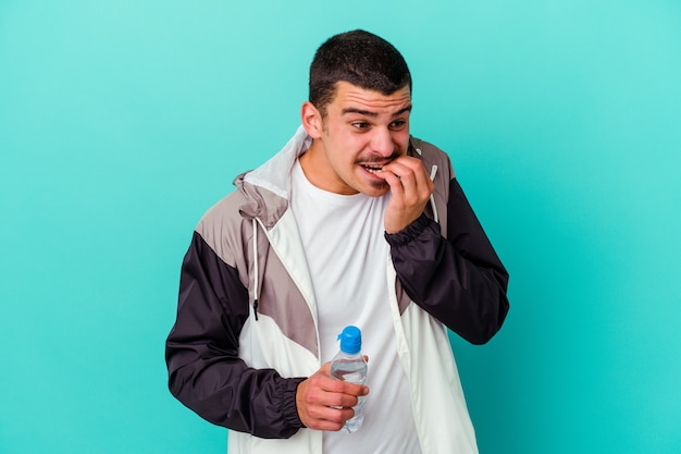 Foto joven deportivo caucásico agua potable aislado en azul mordiéndose las uñas, nervioso y muy ansioso.