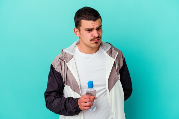 Joven deportivo caucásico beber agua aislado en azul confundido, se siente dudoso e inseguro.