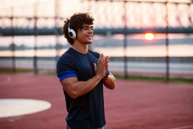 Joven deportivo fuerte con auriculares de pie en el campo de entrenamiento y preparación para el trabajo de la mañana.