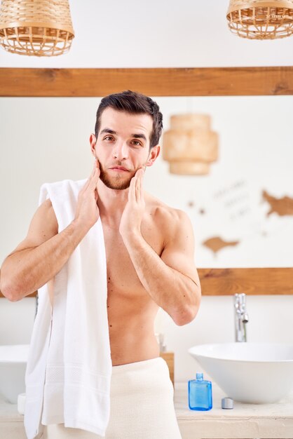 Foto joven deportivo se lava el cuello y se para con una toalla sobre los hombros frente al espejo del baño