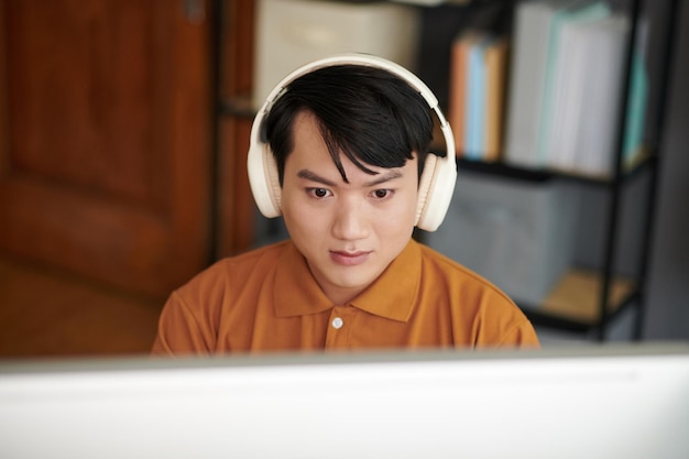 Joven desarrollador de software asiático usando auriculares cuando trabaja en una computadora en la oficina del hogar