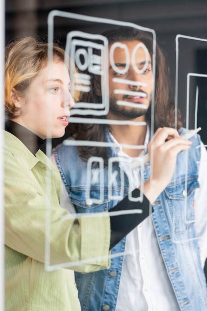 Foto joven desarrollador de software femenino seguro apuntando al boceto de la nueva aplicación móvil mientras hace la presentación a un colega masculino