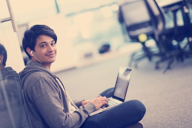 Joven desarrollador de software indio hombre usando una computadora portátil escribiendo código de programación mientras está sentado en el piso en la moderna oficina de inicio creativo