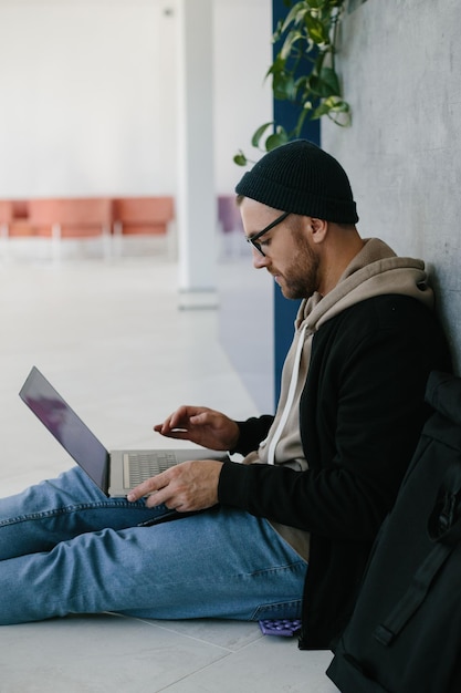 Joven desarrollador de software usando computadora portátil escribiendo código de programación mientras está sentado en el suelo en la moderna oficina creativa de inicio