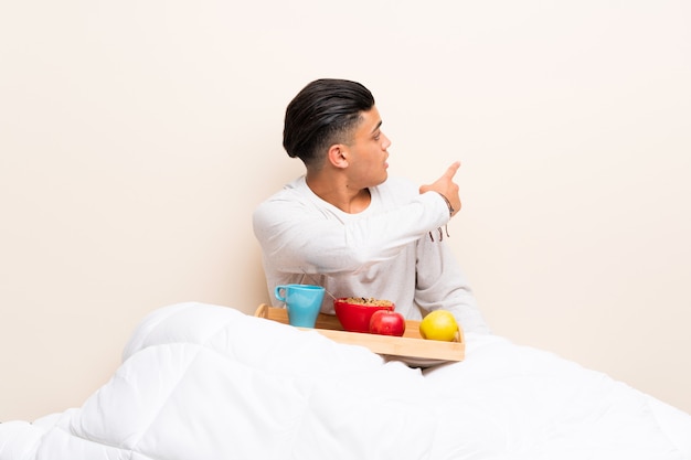 Joven desayunando en la cama apuntando hacia atrás con el dedo índice