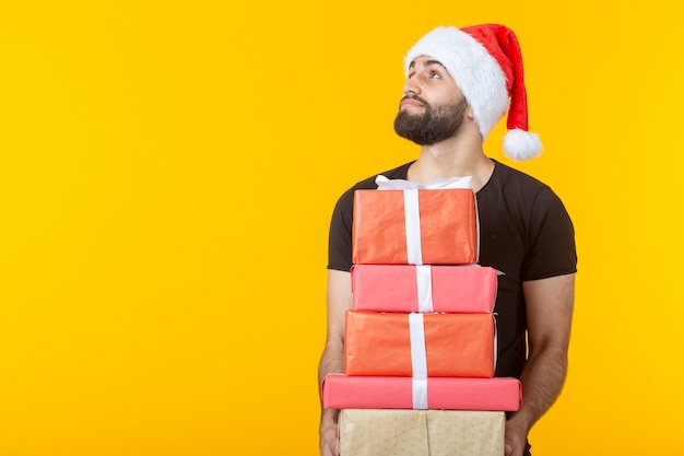 Un joven descontento con barba con un sombrero de Santa Claus tiene cinco cajas de regalo posando sobre un fondo amarillo con copyspace. Concepto de regalos y saludos para Navidad y año nuevo.