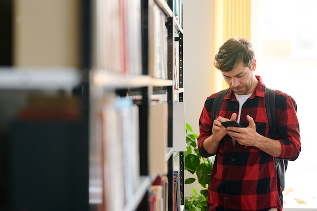 Joven desplazándose en el teléfono inteligente por estante en la biblioteca mientras busca el nombre del libro