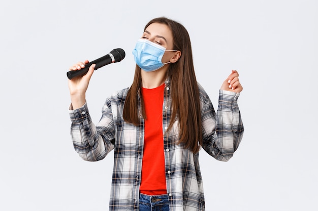 Joven despreocupada en máscara médica, cantando con micrófono, ojos cerrados, disfrutando de karaoke