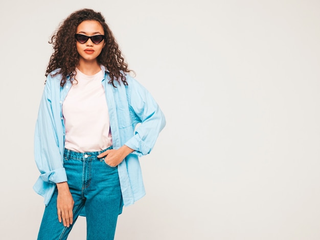 Joven despreocupada posando cerca de azul en estudio con gafas de sol