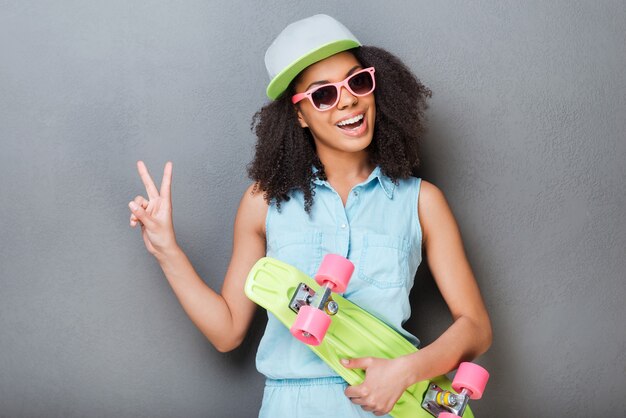 Joven y despreocupado. Atractiva mujer africana joven sosteniendo patineta colorida y sonriendo mientras está de pie contra el fondo gris