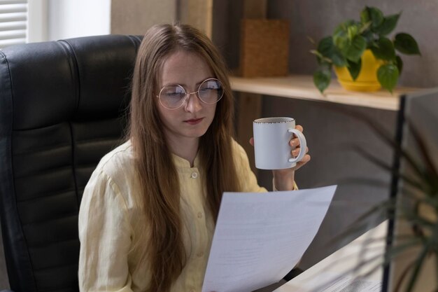 Joven director con gafas está sentado en una silla, tomando café y leyendo documentos. La jefa revisa los informes anuales.