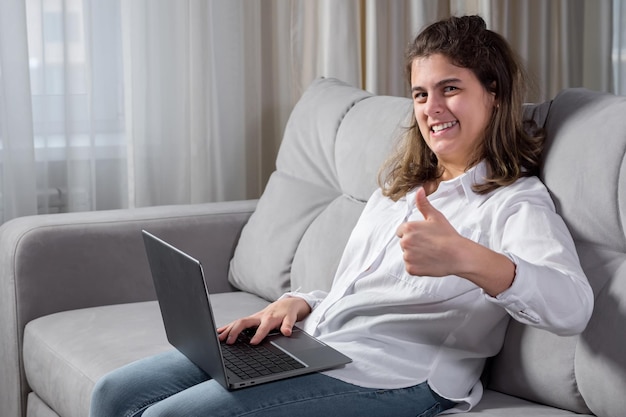 Una joven discapacitada con parálisis cerebral trabaja de forma remota en una computadora tratando de escribir texto con dedos poco móviles sentados en un sofá junto a la ventana en casa