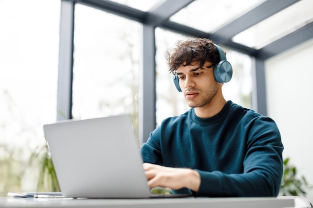 Joven diseñador europeo escuchando música en auriculares mientras trabaja en una computadora portátil en un escritorio en