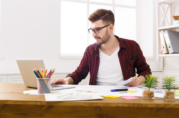 Joven diseñador serio en el trabajo informal con planos y paleta de colores en el interior del lugar de trabajo moderno, espacio de copia