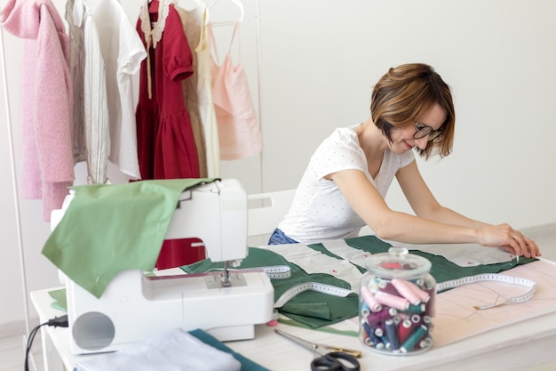 Joven diseñadora hace marcas para un nuevo producto de costura sentado en la mesa junto a la máquina de coser. Concepto de diseño y negocios creativos.