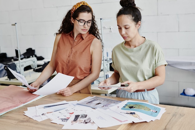 Joven diseñadora mirando la pantalla de la tableta digital en poder de su colega mientras elige modelos para la nueva colección de ropa de temporada