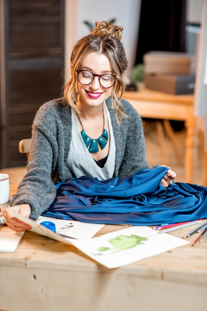 Joven diseñadora de moda trabajando con tela azul sentado en la hermosa oficina con diferentes herramientas de confección en la mesa