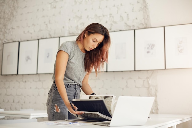 Joven diseñadora de ropa hojeando una revista usando una computadora portátil para encontrar textiles de moda