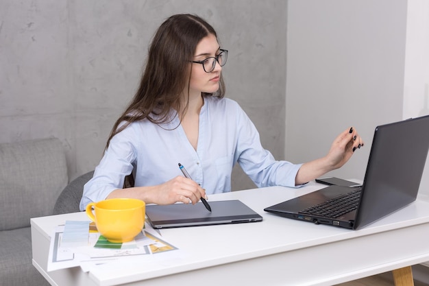 Una joven diseñadora sentada en una mesa dibuja en una tableta. Hace cálculos comerciales en la computadora.