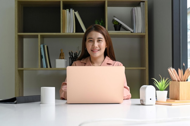 Joven diseñadora sentada en la oficina con su computadora portátil y sonriendo a la cámara.