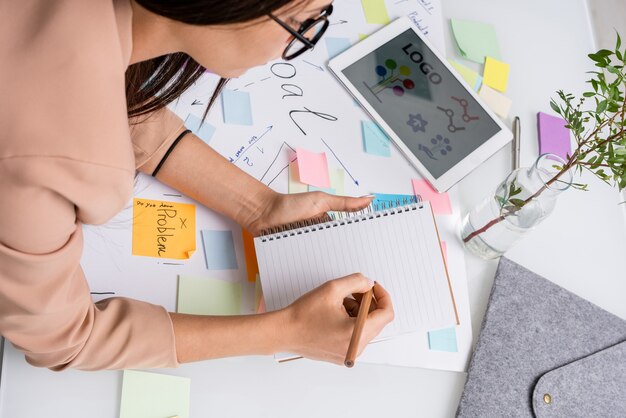 Foto joven diseñadora tomando notas en un cuaderno mientras se inclina sobre un documento grande con objetivos principales y una tableta con un nuevo logotipo en la mesa