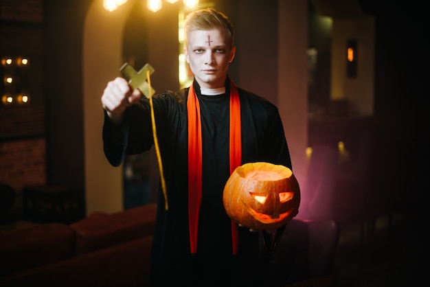 Joven disfrazado de sacerdote de Halloween sostiene una calabaza tallada festiva y una gran cruz negra religiosa. El chico en forma de sacerdote mira a la cámara.