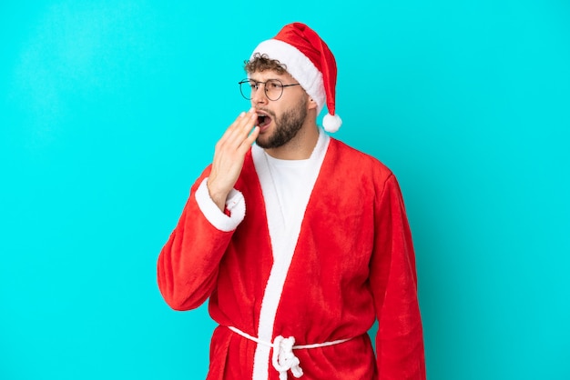 Joven disfrazado de Santa Claus aislado sobre fondo azul bostezando y cubriendo la boca abierta con la mano