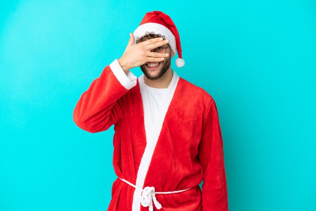Joven disfrazado de Santa Claus aislado sobre fondo azul cubriendo los ojos con las manos y sonriendo