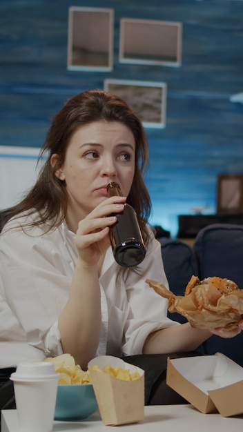 Joven disfrutando de una hamburguesa para llevar y una cerveza después del trabajo sentado en casa en el sofá. mujer comiendo comida rápida y bebiendo bebidas alcohólicas mientras ve la televisión en la sala de estar