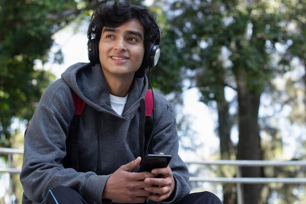 Joven disfrutando de un tiempo solo relajándose escuchando música con sus auriculares en el parque, tiene una sonrisa en la cara y una expresión facial de paz y bienestar.