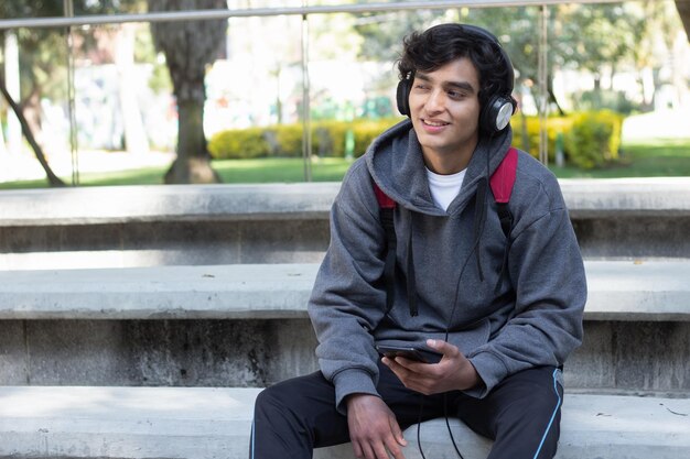 Joven disfrutando de un tiempo solo relajándose escuchando música con sus auriculares en el parque, tiene una sonrisa en la cara y una expresión facial de paz y bienestar.