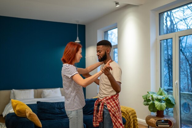 Una joven y diversa pareja de casados alegres bailando y divirtiéndose en la sala de estar de su casa.