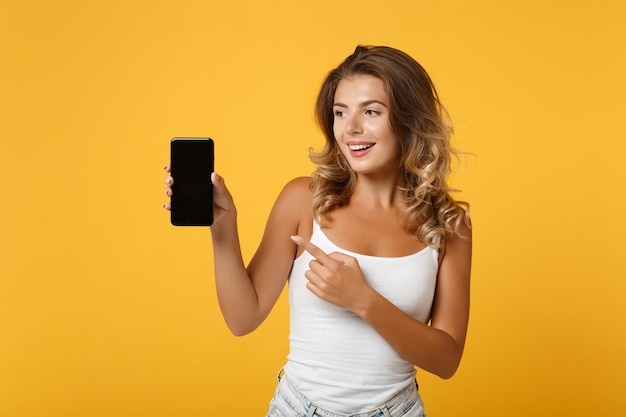 Una joven divertida con ropa ligera e informal posando aislada en un fondo amarillo anaranjado. Concepto de estilo de vida de las personas. Simulacros de espacio de copia. Señalar con el dedo índice en el teléfono móvil con pantalla vacía en blanco.