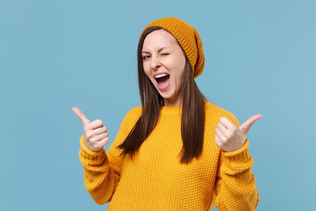 Una joven divertida con suéter amarillo y sombrero posando aislada en un retrato de estudio de fondo de pared azul. Gente emociones sinceras concepto de estilo de vida. Simulacros de espacio de copia. Parpadeando mostrando los pulgares hacia arriba.