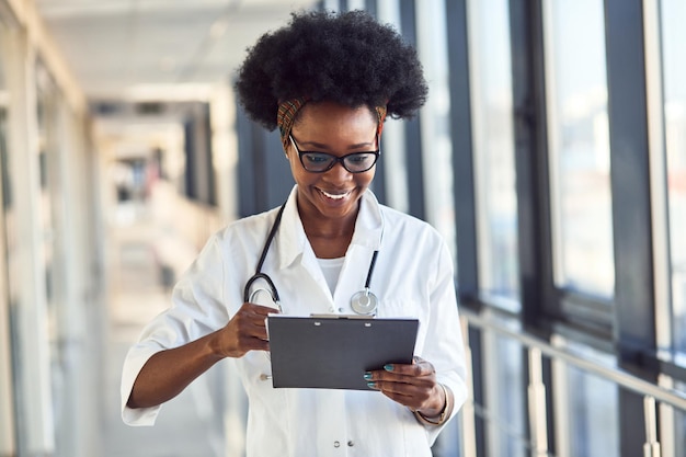 Joven doctora afroamericana en uniforme blanco con estetoscopio y libreta de pie en el pasillo.