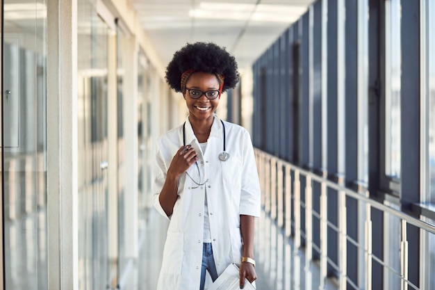 Joven doctora afroamericana en uniforme blanco con estetoscopio y libreta de pie en el pasillo.