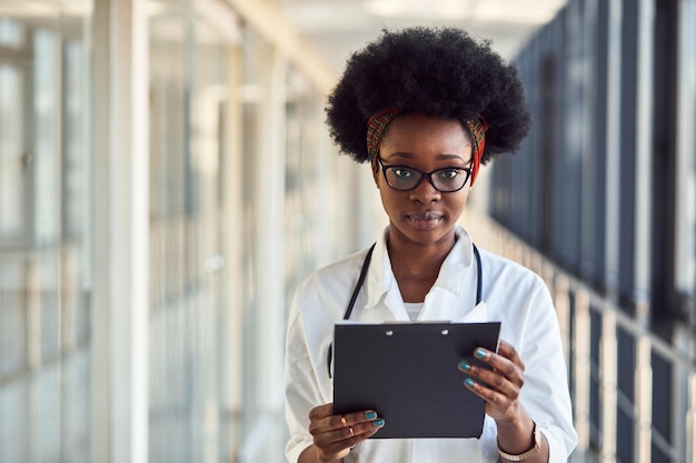 Joven doctora afroamericana en uniforme blanco con estetoscopio y libreta de pie en el pasillo.