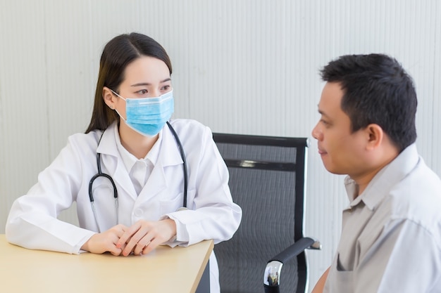 Joven doctora asiática que hace preguntas al paciente en la sala de examen clínico del hospital usando una máscara quirúrgica en todo momento en el concepto de atención médica y protección contra el coronavirus.