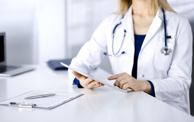 Una joven doctora desconocida está revisando algunos nombres de medicamentos, mientras está sentada en el escritorio de su gabinete en una clínica. El médico con un estetoscopio está usando una tableta, primer plano. médico perfecto s