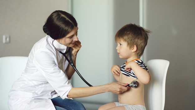 Joven doctora escuchando a un niño pequeño con estetoscopio en el consultorio médico
