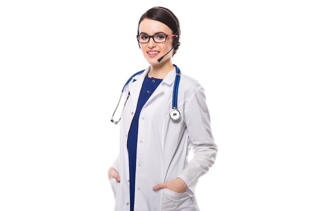 Joven doctora con estetoscopio y auriculares sosteniendo sus manos en los bolsillos en uniforme blanco