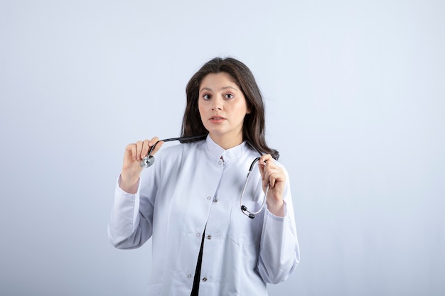 Joven doctora con estetoscopio posando contra la pared blanca.