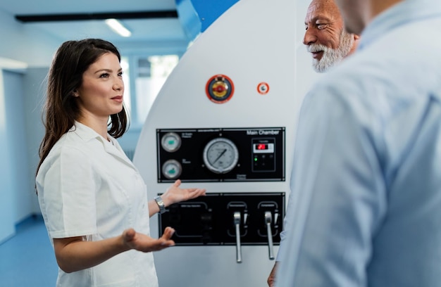 Joven doctora feliz comunicándose con sus pacientes frente a la cámara hiperbárica y explicándoles los beneficios para la salud de la oxigenoterapia