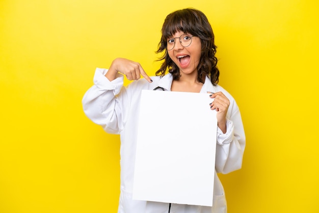 Joven doctora latina aislada de fondo amarillo sosteniendo un cartel vacío con expresión feliz y apuntándolo