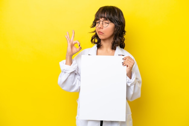 Joven doctora latina aislada de fondo amarillo sosteniendo un cartel vacío y haciendo el signo de OK