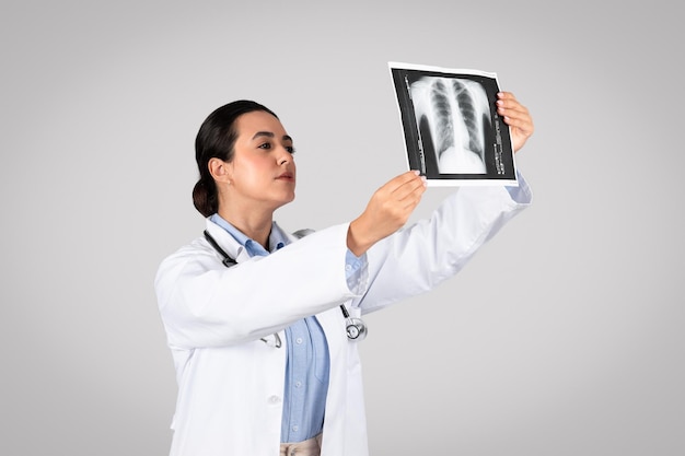 Joven doctora latina sosteniendo y mirando la radiografía de tórax de pie sobre fondo gris del estudio